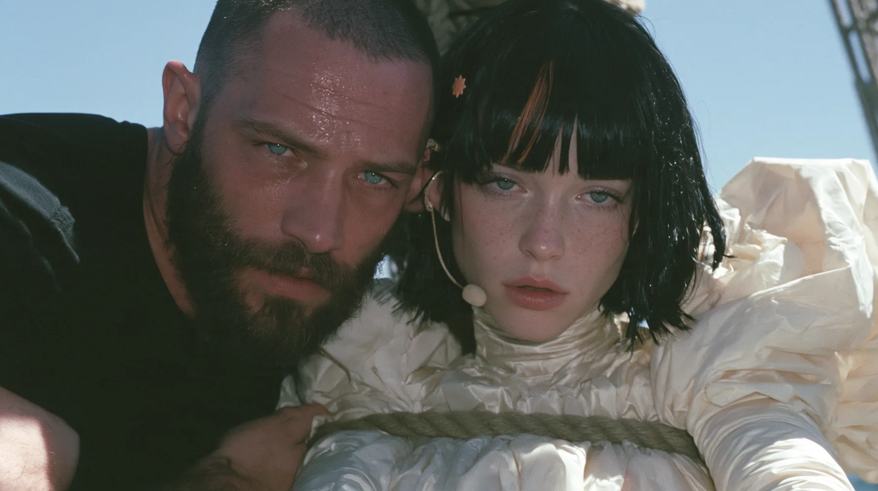 close-up of a broad-shouldered Caucasian man in his early thirties with a shaved head, a thick short dark beard, a square jaw, heavy brows and pale blue eyes, the build of a man who lifts, in a plain black t-shirt…