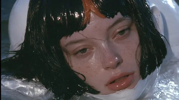 close-up of a colossal smooth white porcelain mask of a young woman's face, sculpted with a glossy black blunt bob with heavy bangs and one clay-orange streak, pale freckles, a serene expression, a hairline crack…