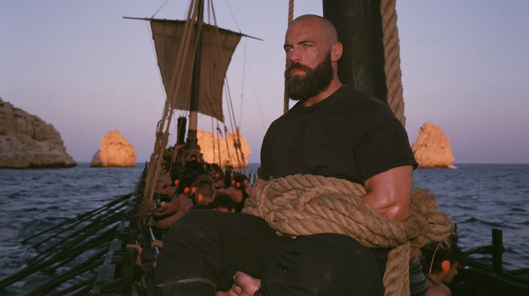 medium wide shot of a broad-shouldered Caucasian man in his early thirties with a shaved head, a thick short dark beard, a square jaw, heavy brows and pale blue eyes, the build of a man who lifts, in a plain black…