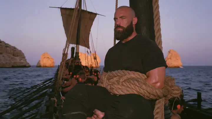medium wide shot of a broad-shouldered Caucasian man in his early thirties with a shaved head, a thick short dark beard, a square jaw, heavy brows and pale blue eyes, the build of a man who lifts, in a plain black…