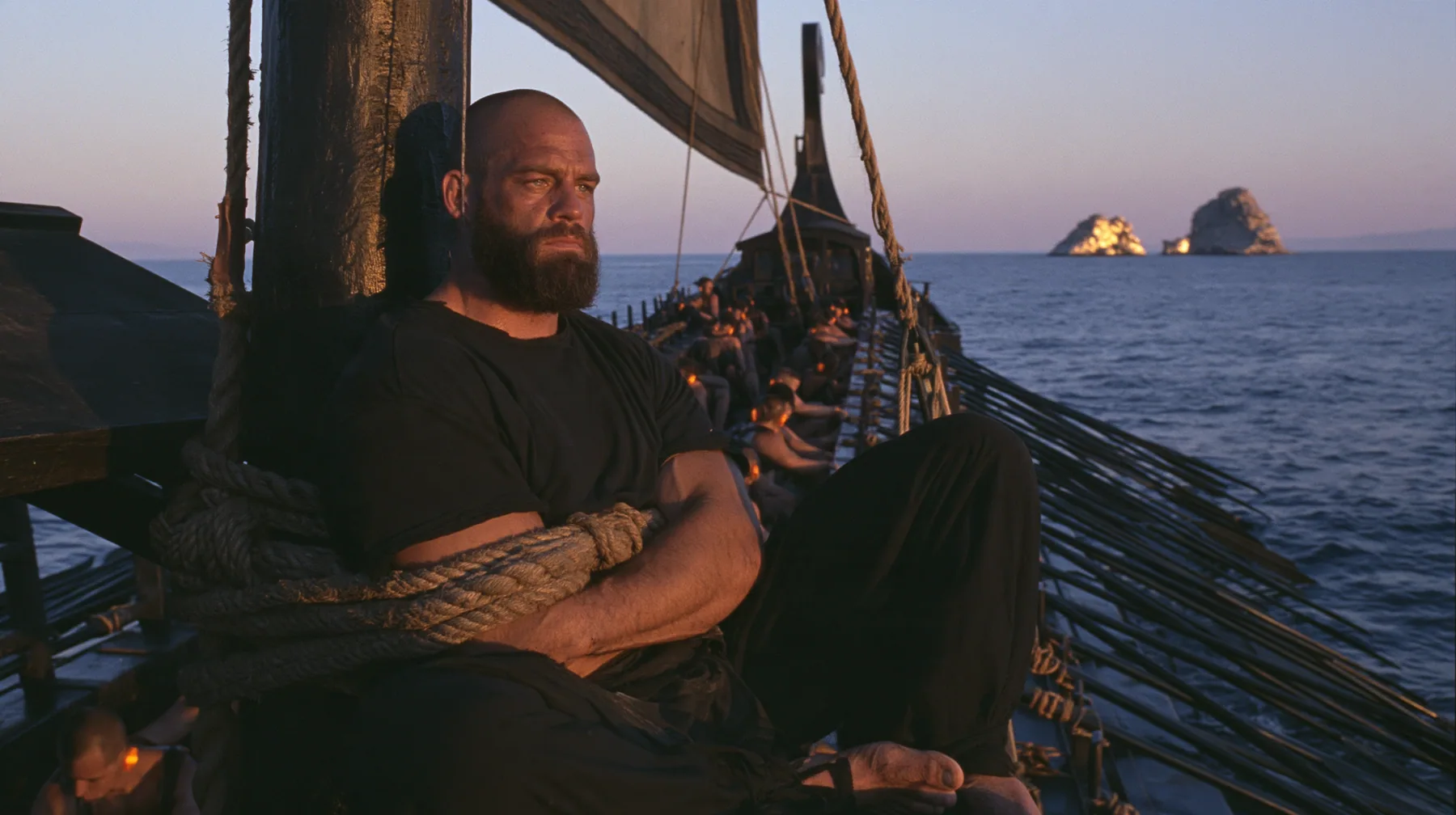 medium wide shot of a broad-shouldered Caucasian man in his early thirties with a shaved head, a thick short dark beard, a square jaw, heavy brows and pale blue eyes, the build of a man who lifts, in a plain black…