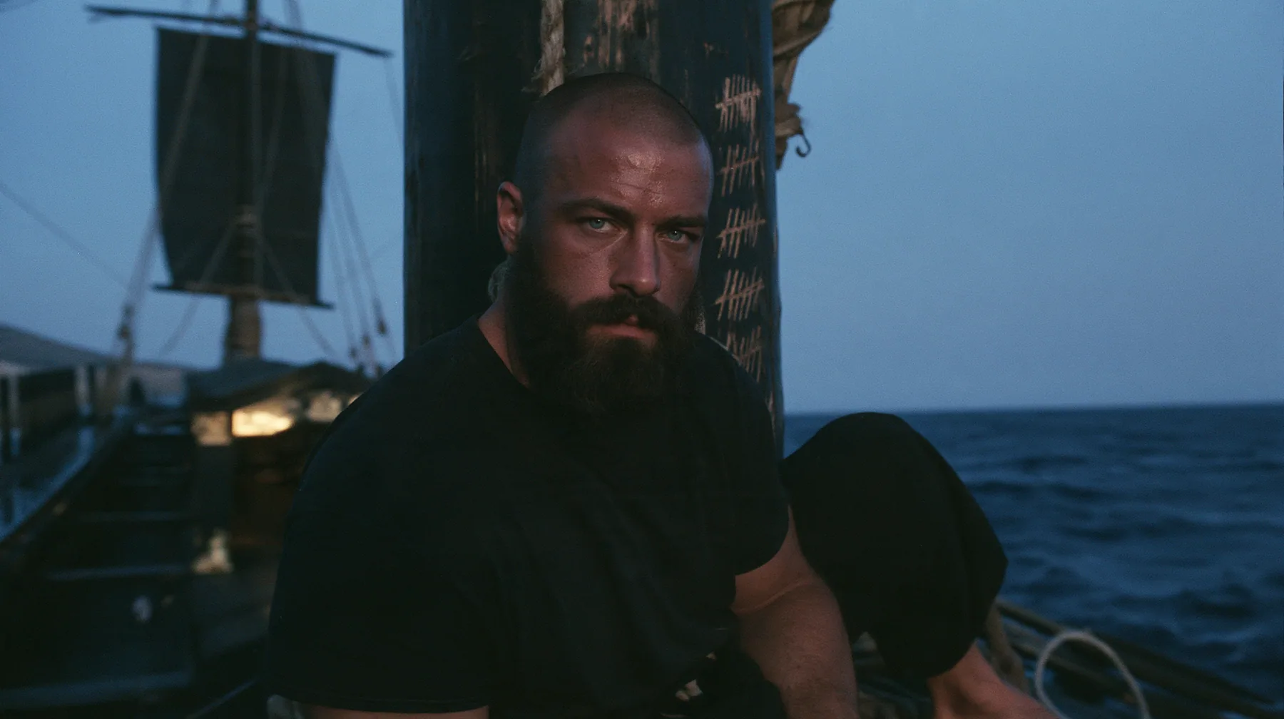 close-up of a broad-shouldered Caucasian man in his early thirties with a shaved head, a thick short dark beard, a square jaw, heavy brows and pale blue eyes, the build of a man who lifts, in a plain black t-shirt…