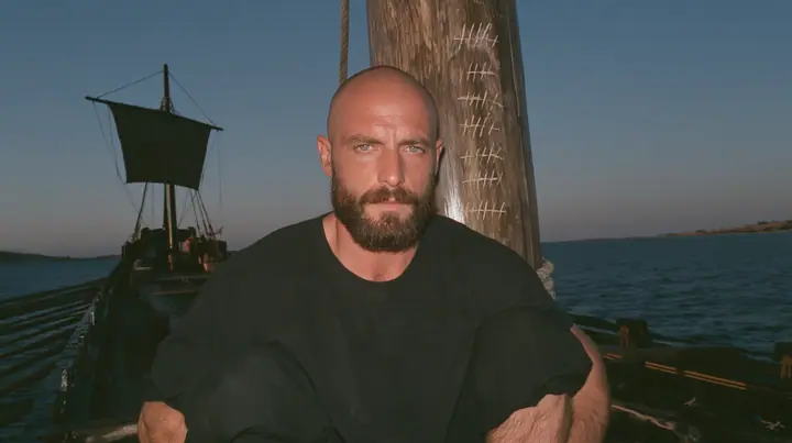 close-up of a broad-shouldered Caucasian man in his early thirties with a shaved head, a thick short dark beard, a square jaw, heavy brows and pale blue eyes, the build of a man who lifts, in a plain black t-shirt…