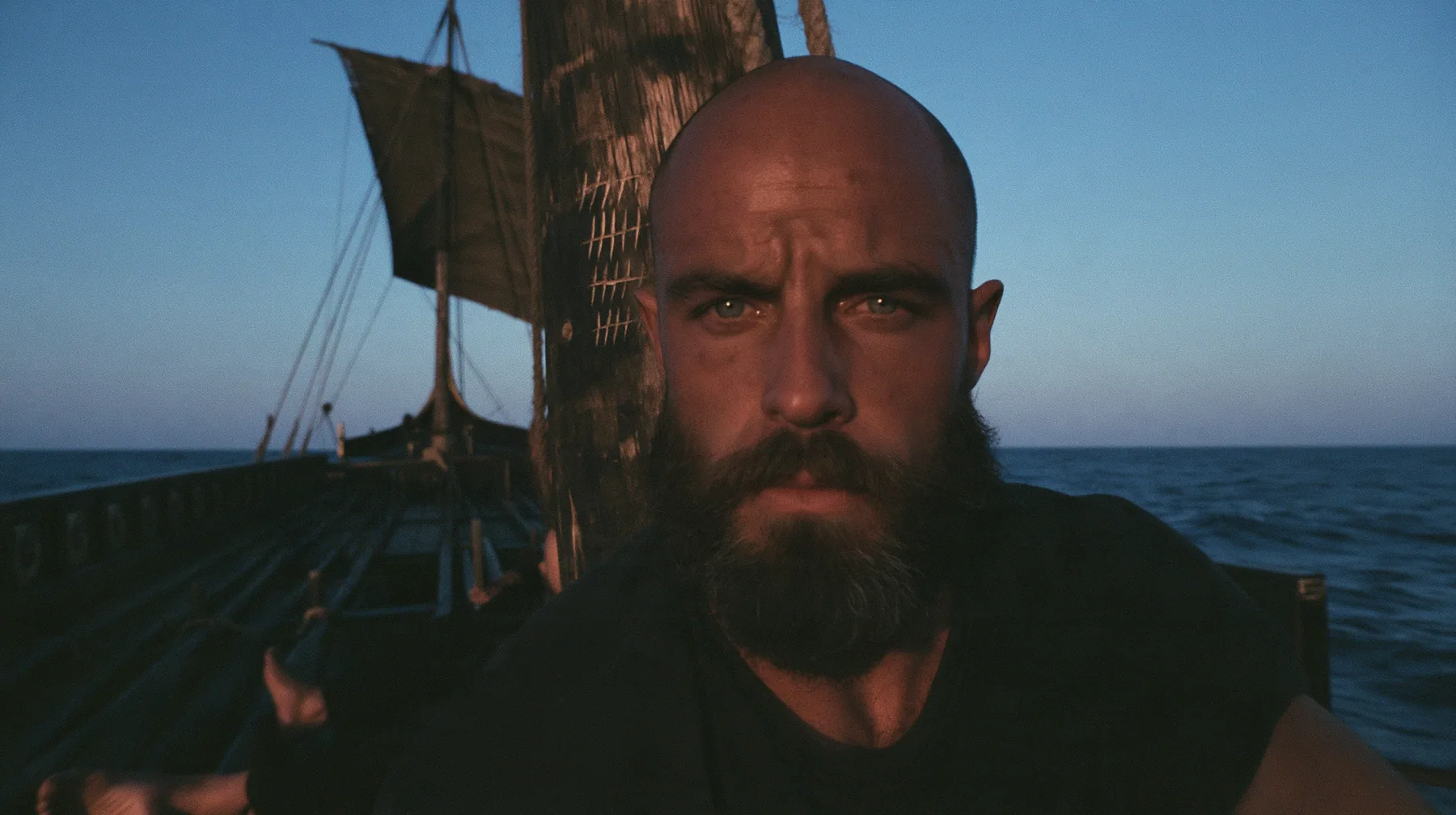 close-up of a broad-shouldered Caucasian man in his early thirties with a shaved head, a thick short dark beard, a square jaw, heavy brows and pale blue eyes, the build of a man who lifts, in a plain black t-shirt…