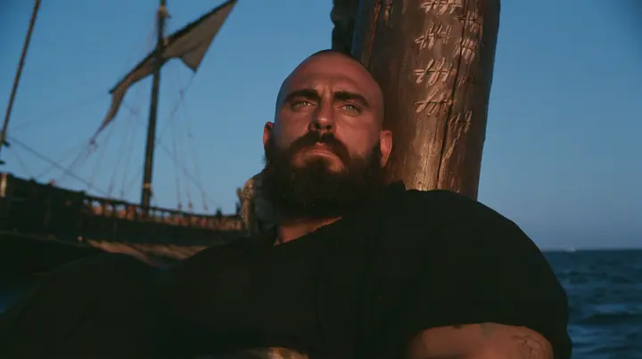 close-up of a broad-shouldered Caucasian man in his early thirties with a shaved head, a thick short dark beard, a square jaw, heavy brows and pale blue eyes, the build of a man who lifts, in a plain black t-shirt…