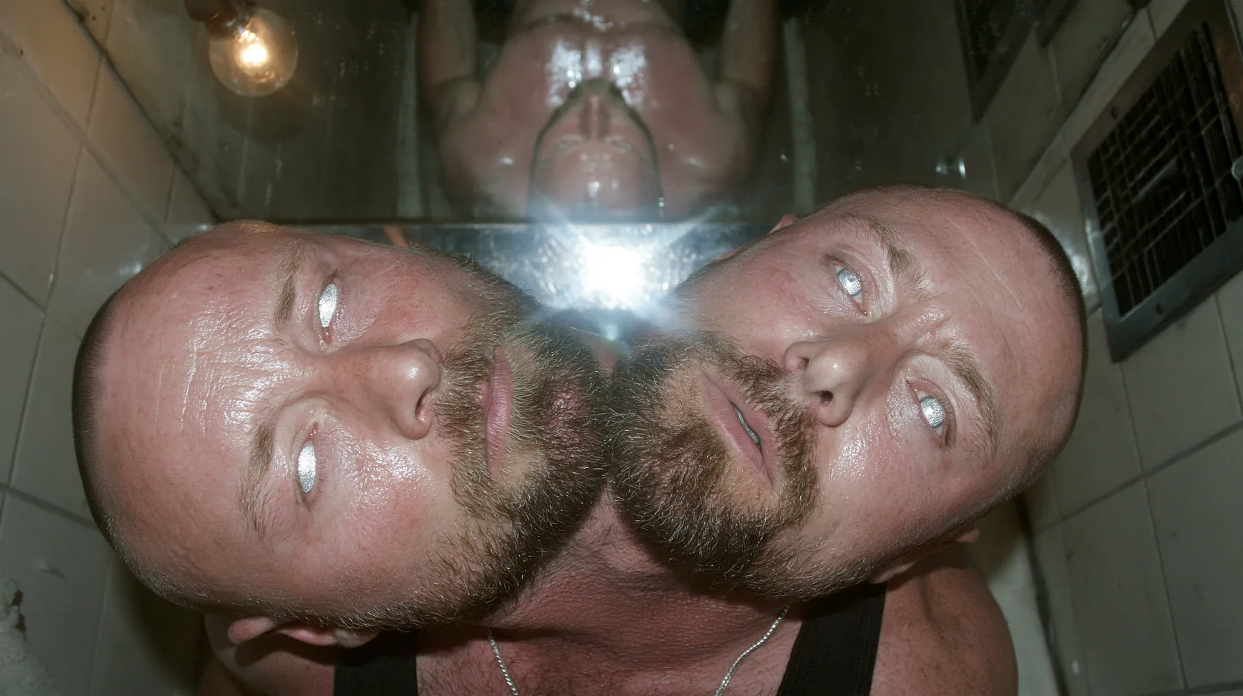 straight-down shot of a broad-shouldered Caucasian man in his early thirties with a shaved head, a thick short dark beard, a square jaw, heavy brows and pale blue eyes, the build of a man who lifts, in a black tank…
