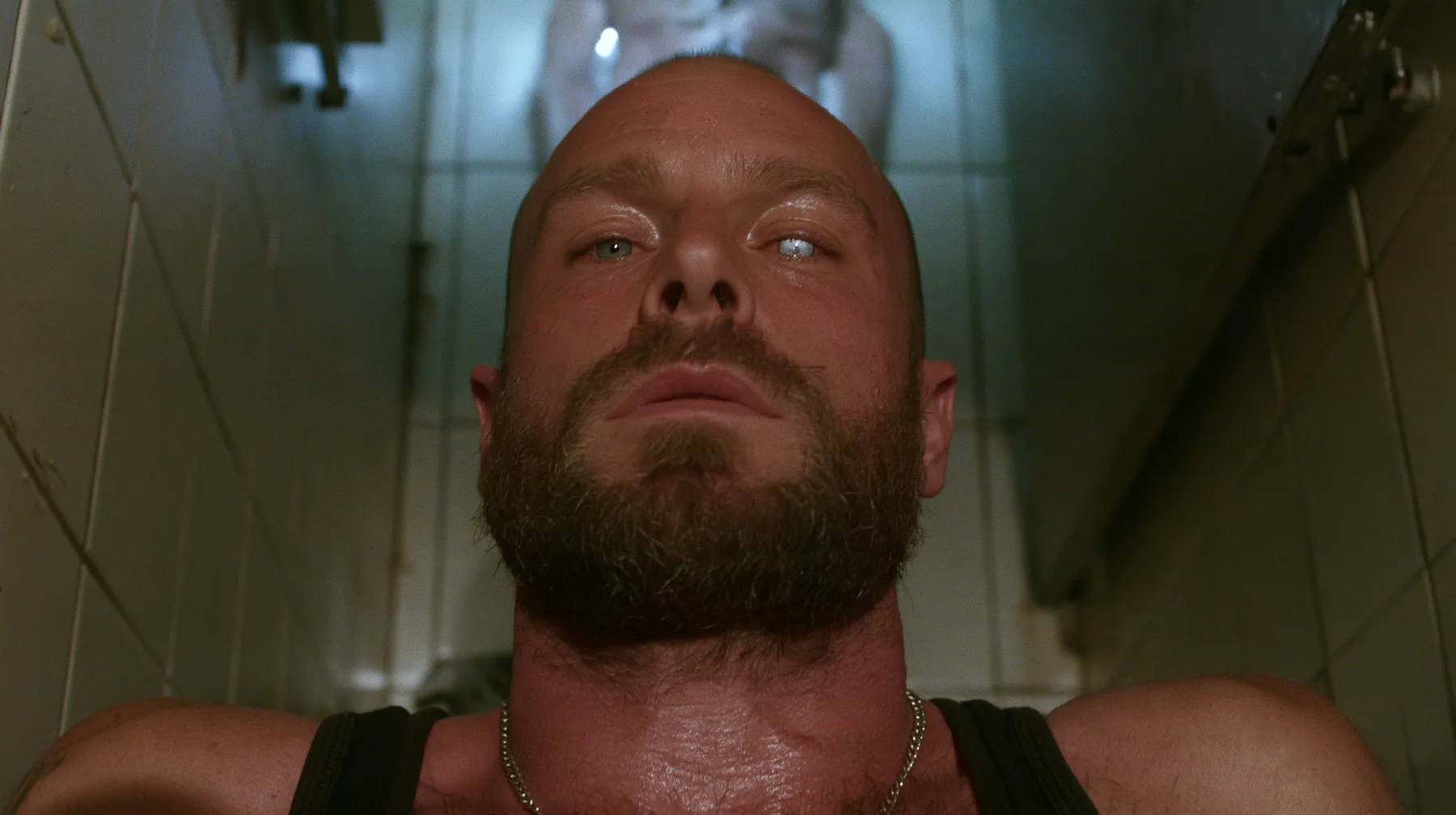 straight-down shot of a broad-shouldered Caucasian man in his early thirties with a shaved head, a thick short dark beard, a square jaw, heavy brows and pale blue eyes, the build of a man who lifts, in a black tank…