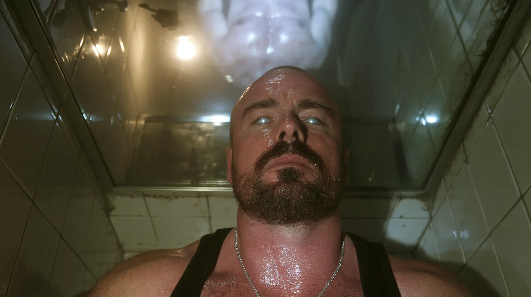 straight-down shot of a broad-shouldered Caucasian man in his early thirties with a shaved head, a thick short dark beard, a square jaw, heavy brows and pale blue eyes, the build of a man who lifts, in a black tank…