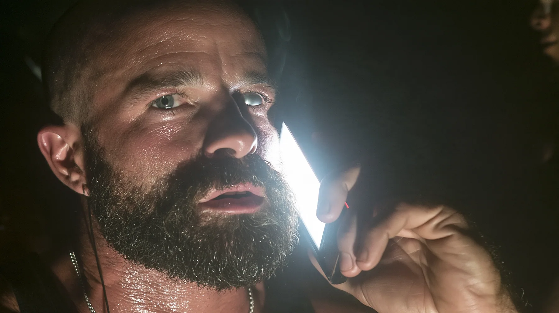 extreme close-up of a broad-shouldered Caucasian man in his early thirties with a shaved head, a thick short dark beard, a square jaw, heavy brows and pale blue eyes, the build of a man who lifts, in a black tank top…