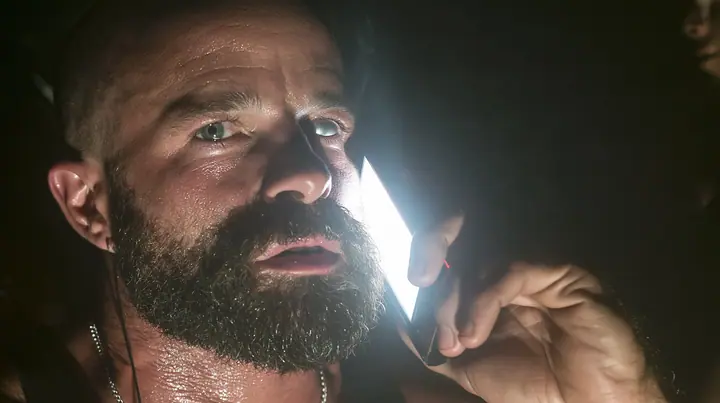 extreme close-up of a broad-shouldered Caucasian man in his early thirties with a shaved head, a thick short dark beard, a square jaw, heavy brows and pale blue eyes, the build of a man who lifts, in a black tank top…