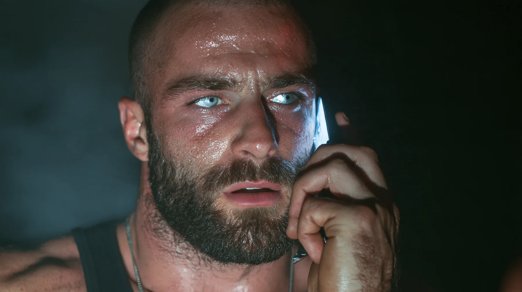 extreme close-up of a broad-shouldered Caucasian man in his early thirties with a shaved head, a thick short dark beard, a square jaw, heavy brows and pale blue eyes, the build of a man who lifts, in a black tank top…