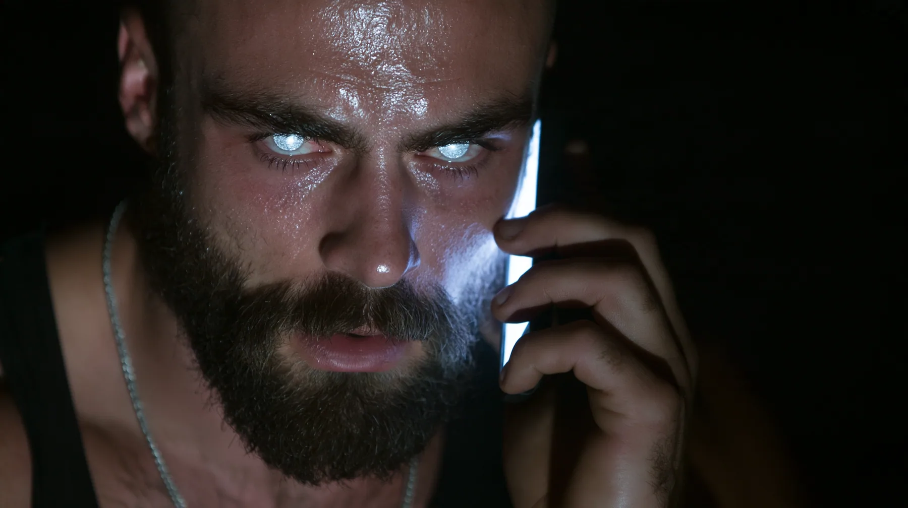 extreme close-up of a broad-shouldered Caucasian man in his early thirties with a shaved head, a thick short dark beard, a square jaw, heavy brows and pale blue eyes, the build of a man who lifts, in a black tank top…