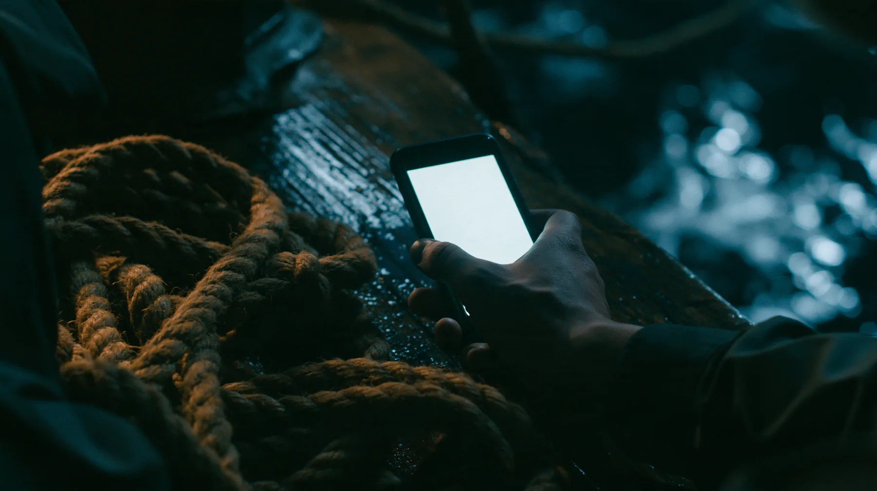 close-up of a man's hand holding a phone on the deck of an ancient ship at night, a coil of thick hemp rope beside it, the phone's screen glowing plain white, moonlight on the wet wood, night, moonlight on a…