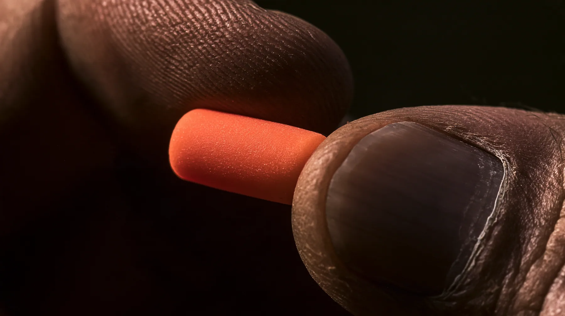 macro of a rower's rough weathered fingers pinching a bright orange foam earplug and rolling it into a thin cylinder between thumb and forefinger, the plug squeezed and held firmly, macro product photograph, hard rim…