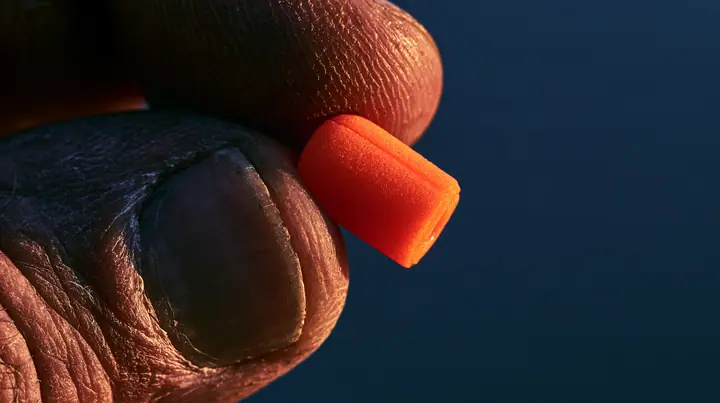 macro of a rower's rough weathered fingers pinching a bright orange foam earplug and rolling it into a thin cylinder between thumb and forefinger, the plug squeezed and held firmly, macro product photograph, hard rim…