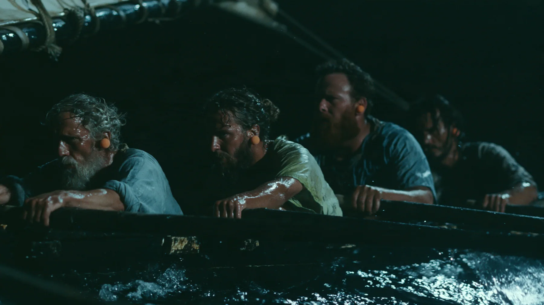 medium shot along a rowing bench of a long black ancient Greek galley with a single mast, a square sail and two banks of oars at night, four oarsmen of different ages and builds pulling their oars together, a…