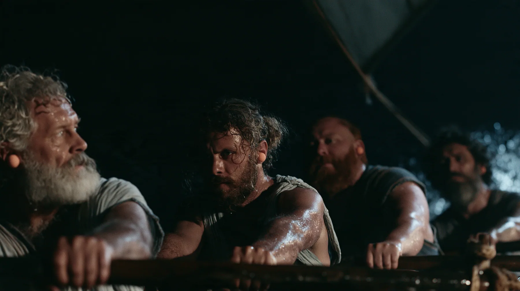 medium shot along a rowing bench of a long black ancient Greek galley with a single mast, a square sail and two banks of oars at night, four oarsmen of different ages and builds pulling their oars together, a…
