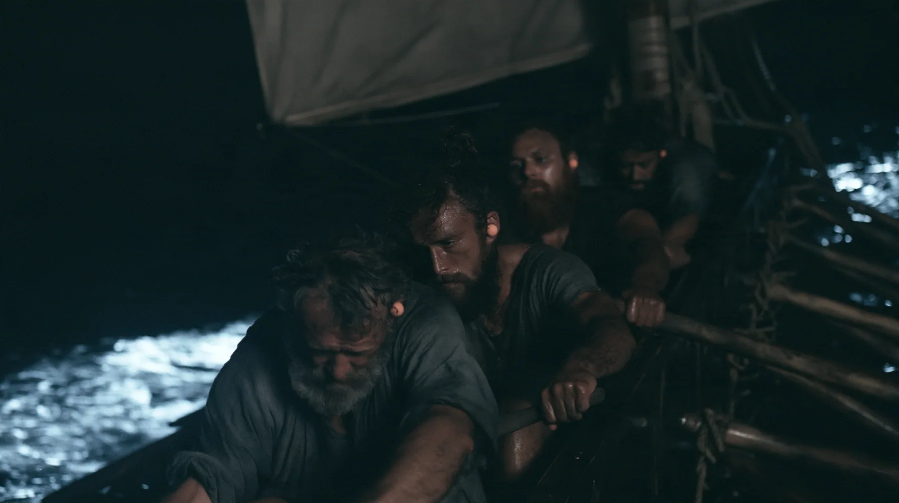 medium shot along a rowing bench of a long black ancient Greek galley with a single mast, a square sail and two banks of oars at night, four oarsmen of different ages and builds pulling their oars together, a…