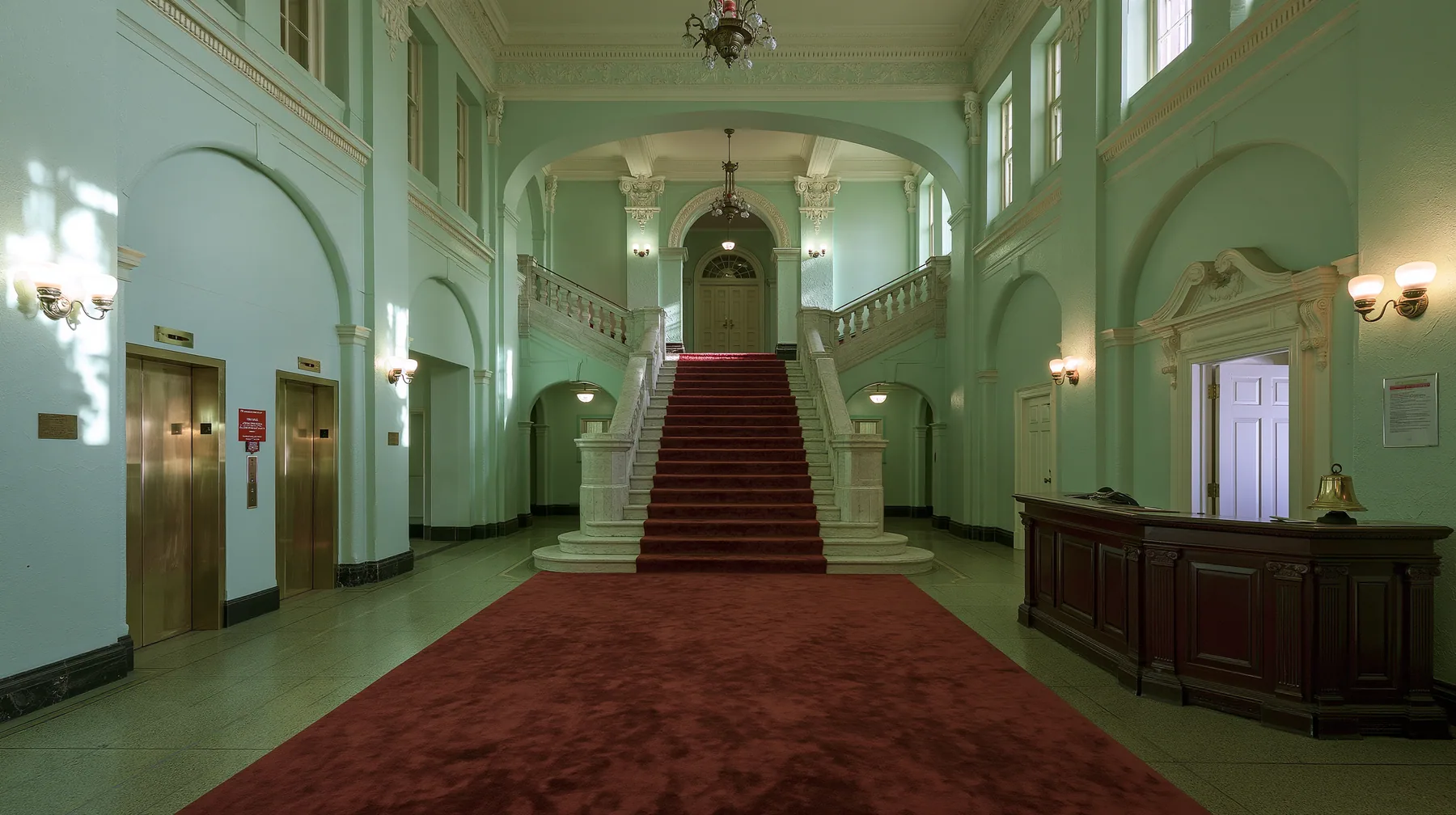 wide perfectly symmetrical interior of a grand hotel lobby: mint-green walls, a red-carpet staircase, brass elevator doors, a mahogany reception desk with a brass bell, warm light, pastel art-house film still, no…