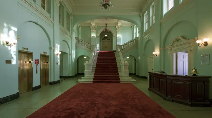 wide perfectly symmetrical interior of a grand hotel lobby: mint-green walls, a red-carpet staircase, brass elevator doors, a mahogany reception desk with a brass bell, warm light, pastel art-house film still, no…