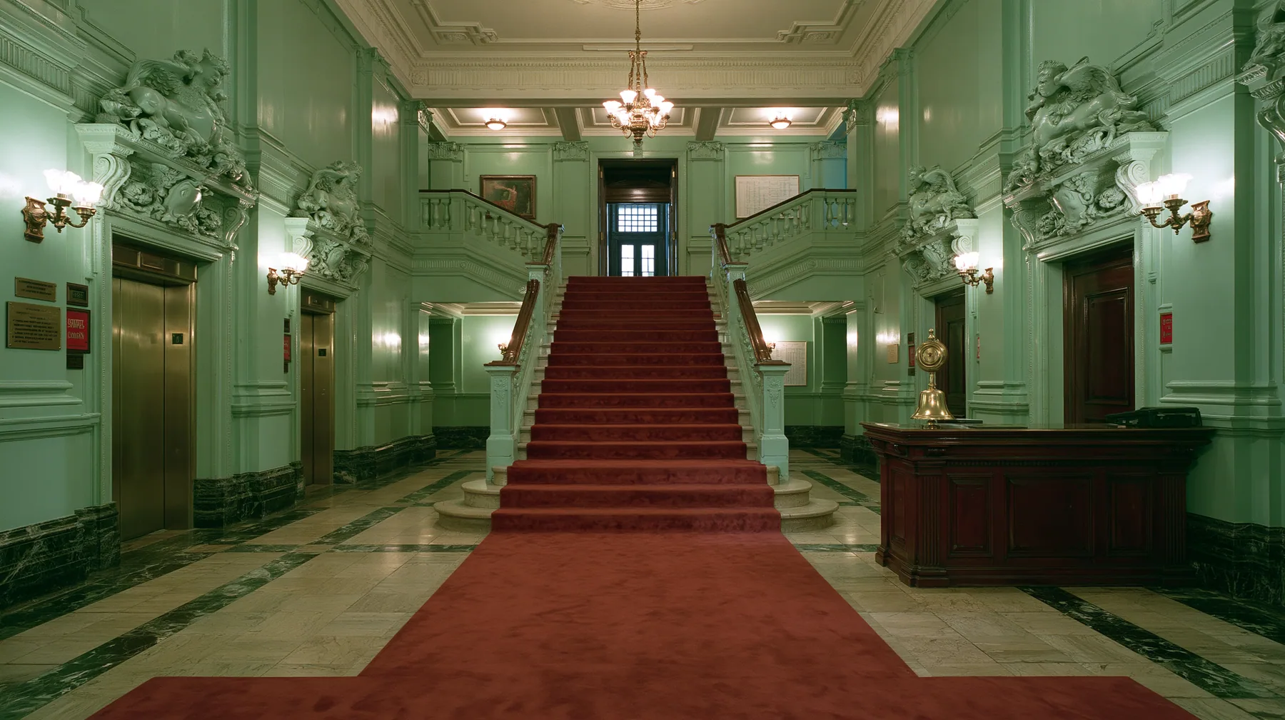 wide perfectly symmetrical interior of a grand hotel lobby: mint-green walls, a red-carpet staircase, brass elevator doors, a mahogany reception desk with a brass bell, warm light, pastel art-house film still, no…