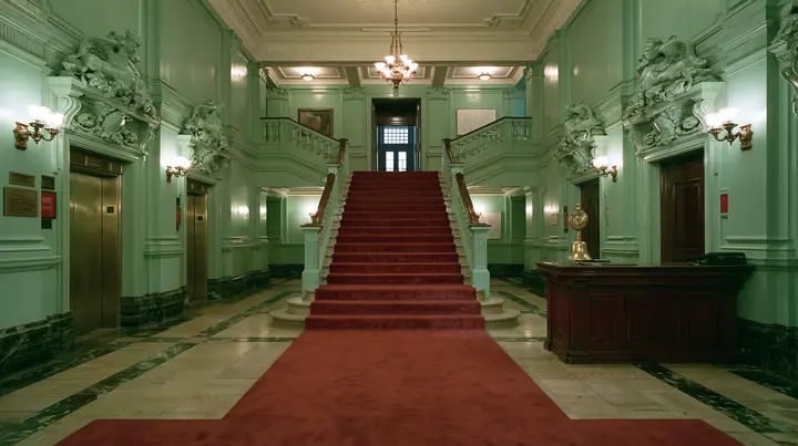 wide perfectly symmetrical interior of a grand hotel lobby: mint-green walls, a red-carpet staircase, brass elevator doors, a mahogany reception desk with a brass bell, warm light, pastel art-house film still, no…