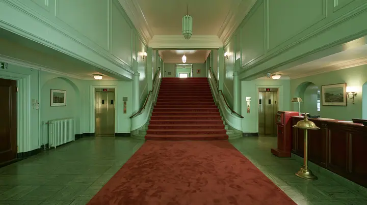 wide perfectly symmetrical interior of a grand hotel lobby: mint-green walls, a red-carpet staircase, brass elevator doors, a mahogany reception desk with a brass bell, warm light, pastel art-house film still, no…