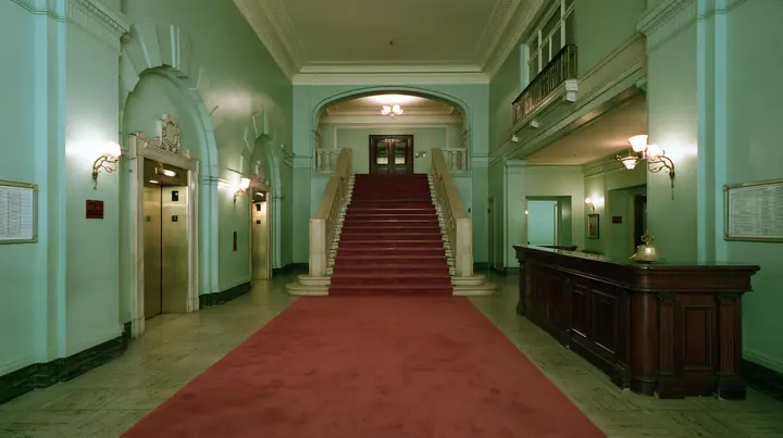 wide perfectly symmetrical interior of a grand hotel lobby: mint-green walls, a red-carpet staircase, brass elevator doors, a mahogany reception desk with a brass bell, warm light, pastel art-house film still, no…