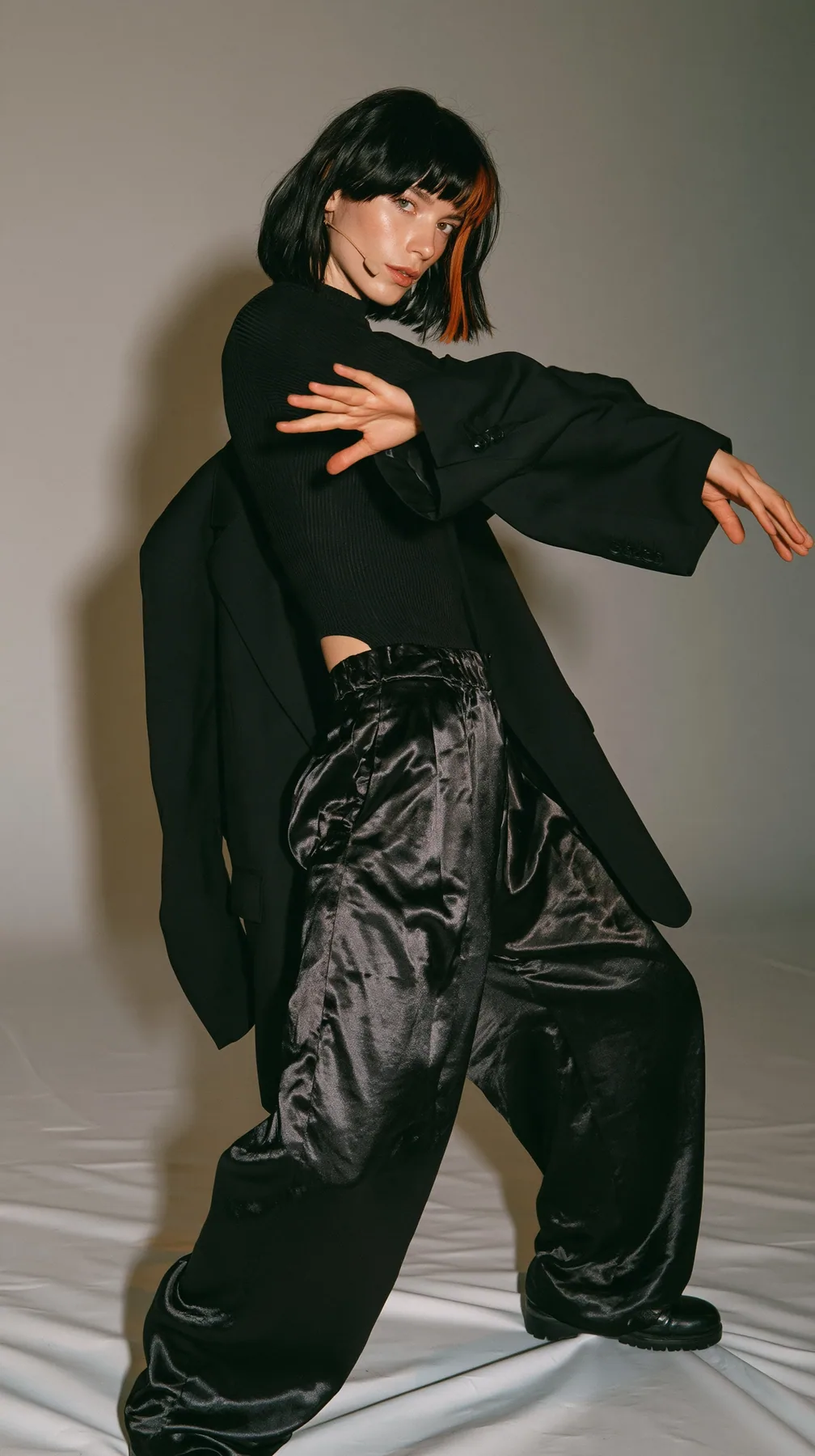 full-length photo, a Caucasian American woman in her late twenties with pale skin and a glossy black blunt bob with heavy straight bangs and one clay-orange streak, a thin headset microphone, in a black ribbed top,…