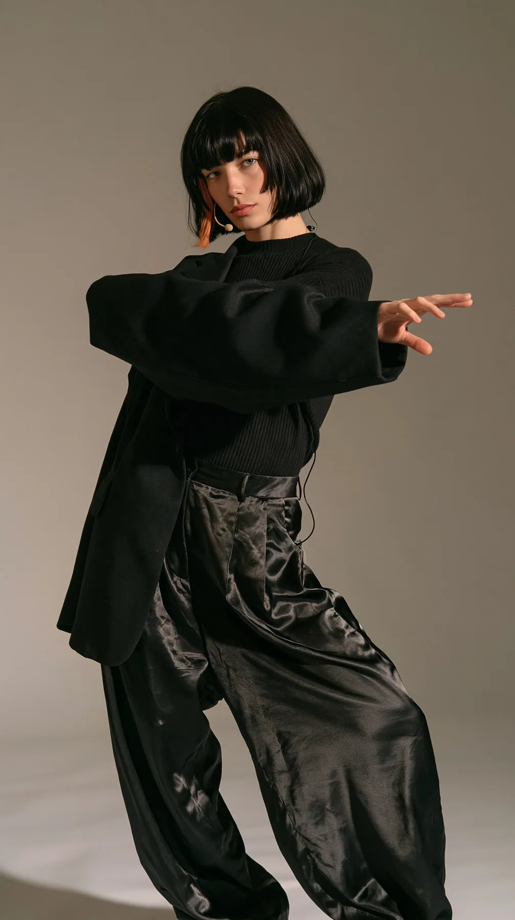 full-length photo, a Caucasian American woman in her late twenties with pale skin and a glossy black blunt bob with heavy straight bangs and one clay-orange streak, a thin headset microphone, in a black ribbed top,…