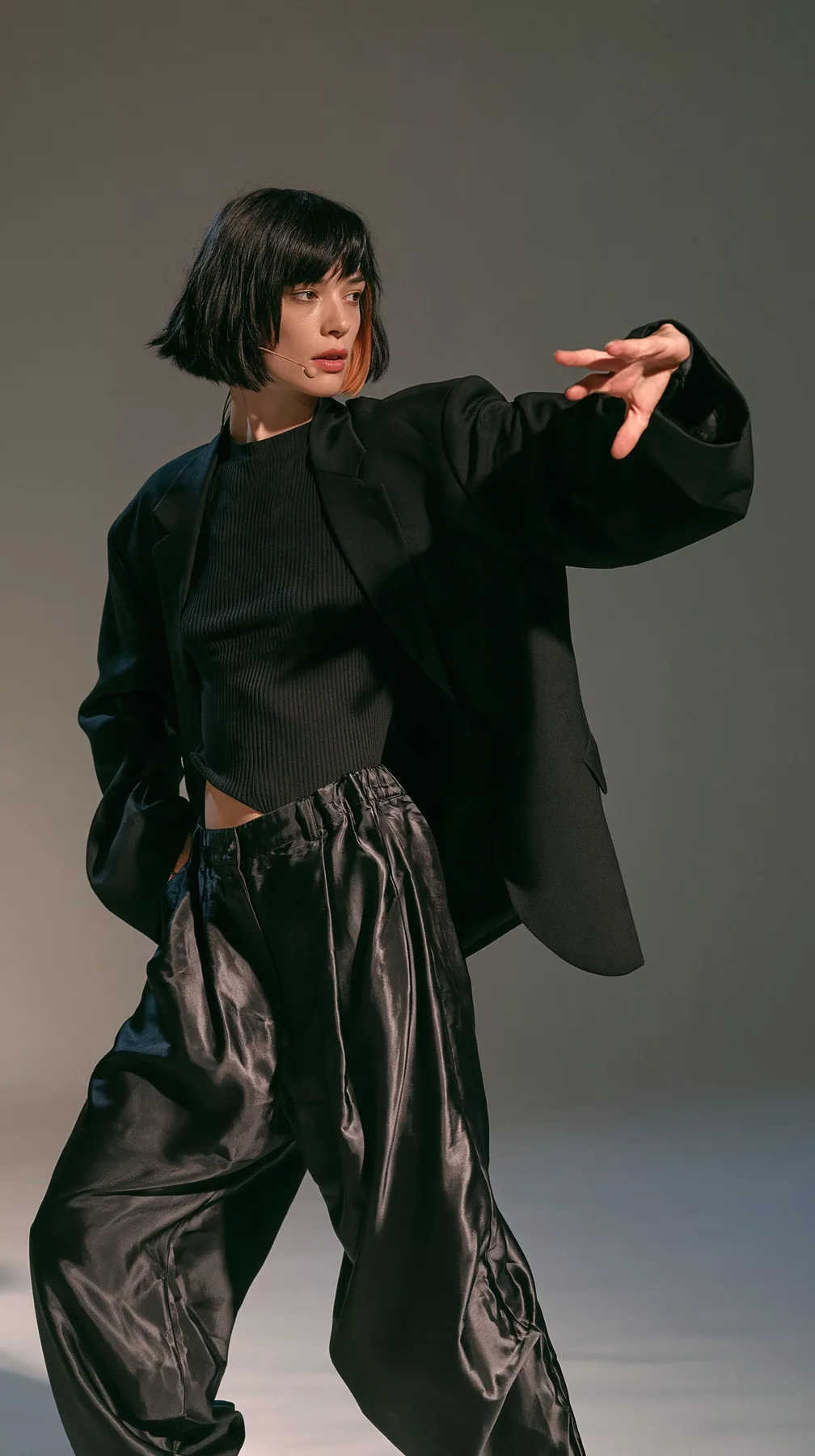full-length photo, a Caucasian American woman in her late twenties with pale skin and a glossy black blunt bob with heavy straight bangs and one clay-orange streak, a thin headset microphone, in a black ribbed top,…