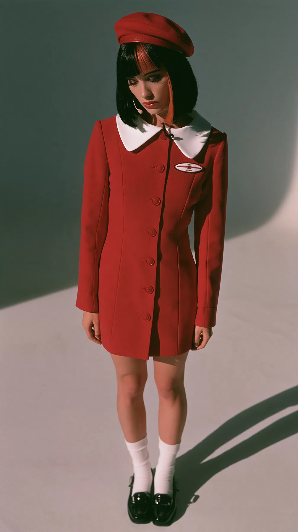 full-length photo from behind, a Caucasian American woman in her late twenties with pale skin and a glossy black blunt bob with heavy straight bangs and one clay-orange streak, a thin headset microphone, in a red…