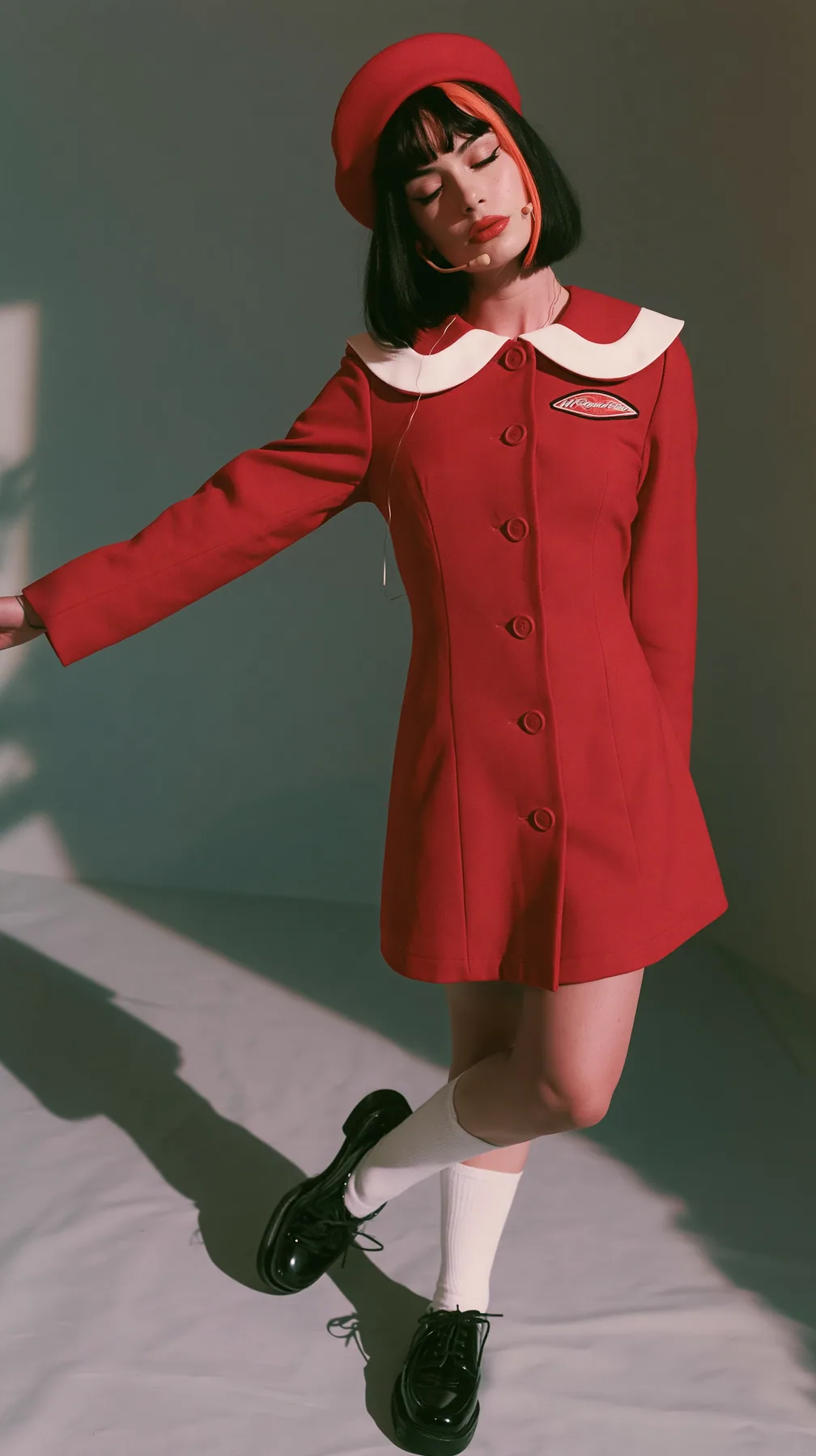 full-length photo, a Caucasian American woman in her late twenties with pale skin and a glossy black blunt bob with heavy straight bangs and one clay-orange streak, a thin headset microphone, in a red tailored coat…