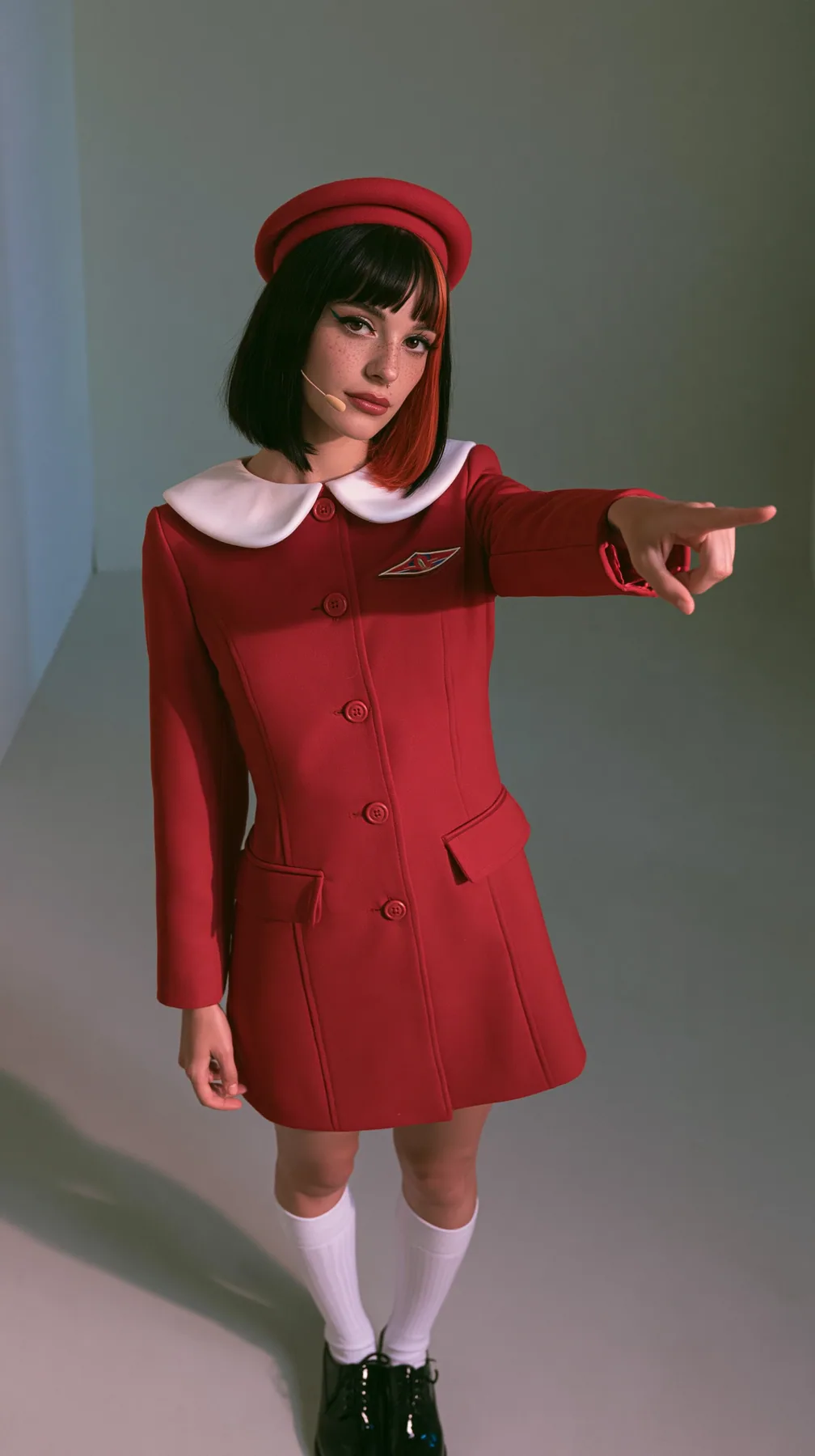 waist-up photo, a Caucasian American woman in her late twenties with pale skin and a glossy black blunt bob with heavy straight bangs and one clay-orange streak, a thin headset microphone, in a red tailored coat…