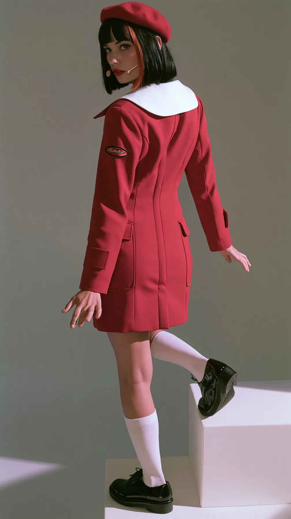 full-length photo from behind, a Caucasian American woman in her late twenties with pale skin and a glossy black blunt bob with heavy straight bangs and one clay-orange streak, a thin headset microphone, in a red…