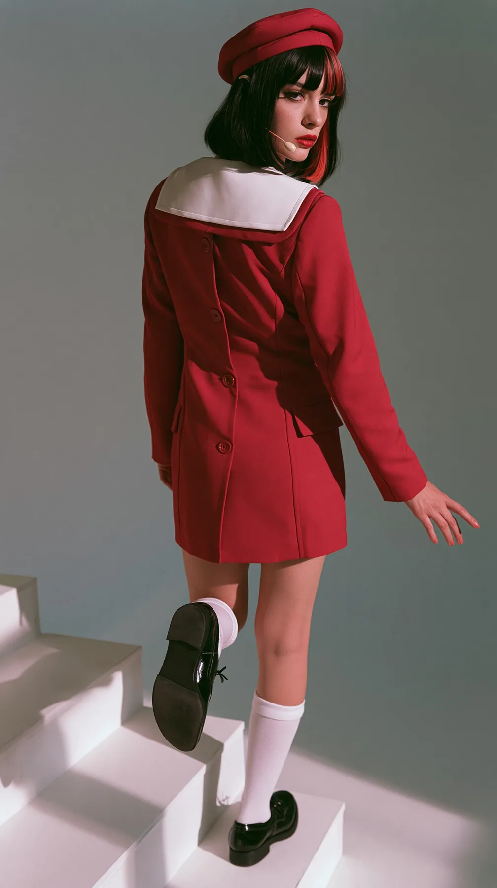 full-length photo from behind, a Caucasian American woman in her late twenties with pale skin and a glossy black blunt bob with heavy straight bangs and one clay-orange streak, a thin headset microphone, in a red…