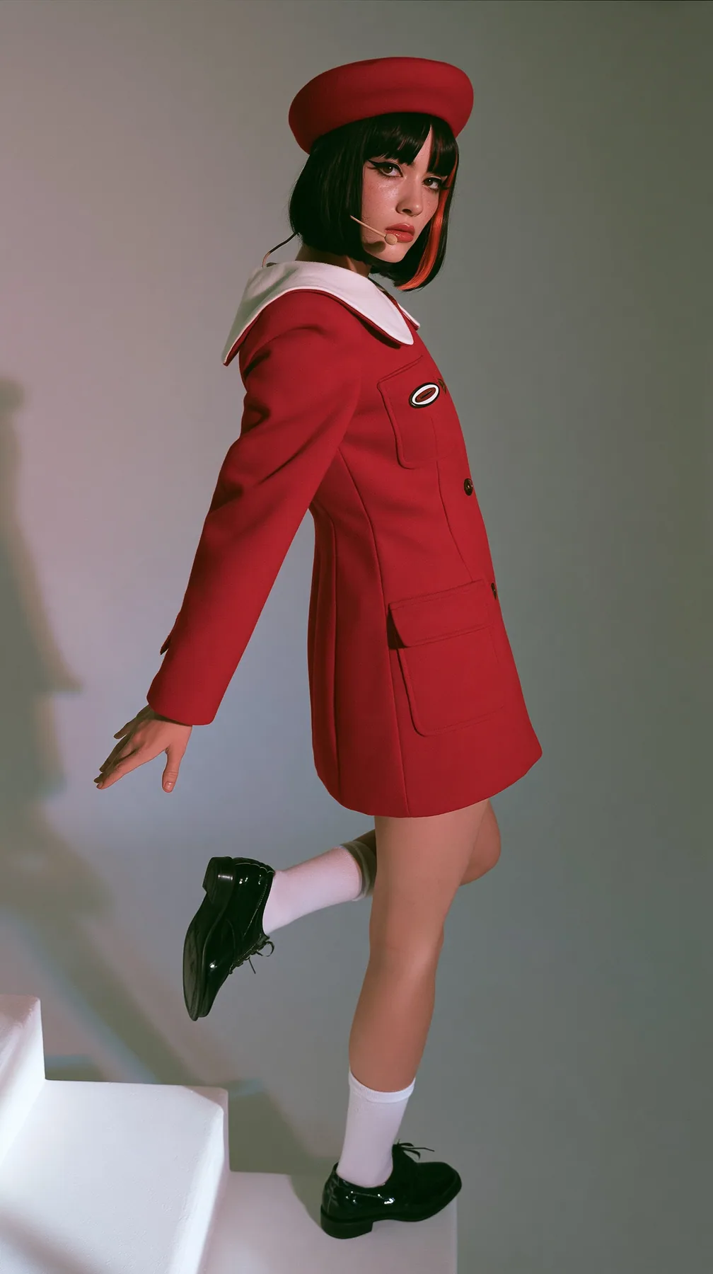 full-length photo from behind, a Caucasian American woman in her late twenties with pale skin and a glossy black blunt bob with heavy straight bangs and one clay-orange streak, a thin headset microphone, in a red…