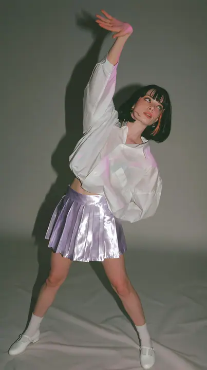 full-length photo, a Caucasian American woman in her late twenties with pale skin and a glossy black blunt bob with heavy straight bangs and one clay-orange streak, a thin headset microphone, in an oversized white…