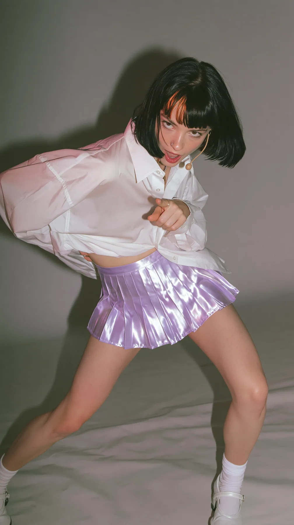 full-length photo, a Caucasian American woman in her late twenties with pale skin and a glossy black blunt bob with heavy straight bangs and one clay-orange streak, a thin headset microphone, in an oversized white…