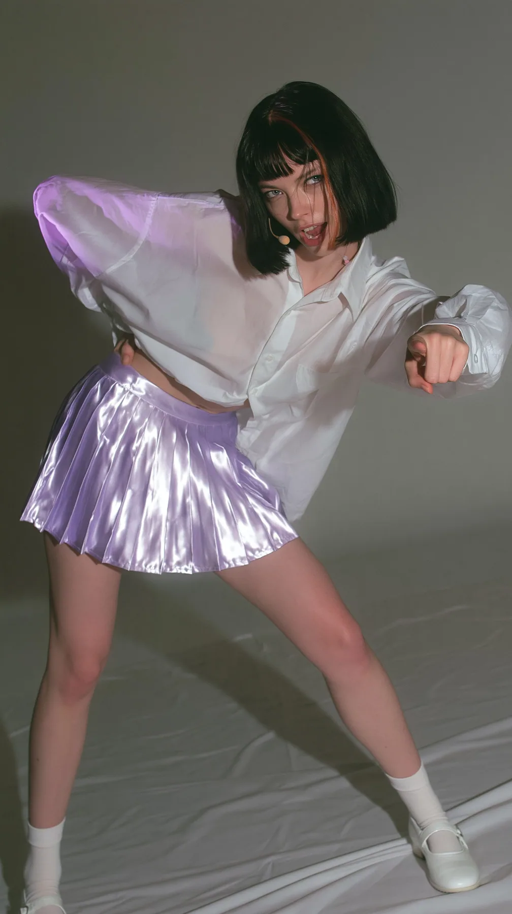 full-length photo, a Caucasian American woman in her late twenties with pale skin and a glossy black blunt bob with heavy straight bangs and one clay-orange streak, a thin headset microphone, in an oversized white…