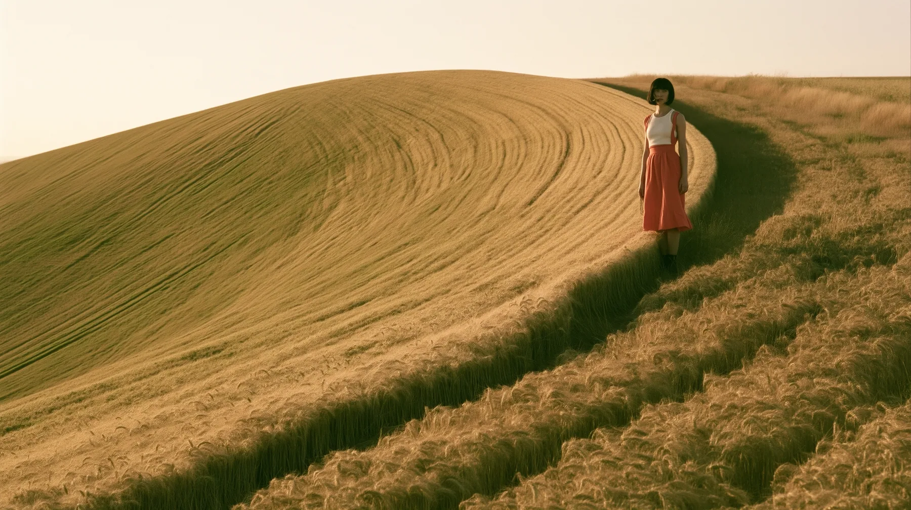 Make this a vast wide 35 mm photograph: a grassy hill shaped exactly like a smooth bell curve rises out of a wheat field, with mown stripes down its slopes; keep this woman, a Caucasian American woman in her late…