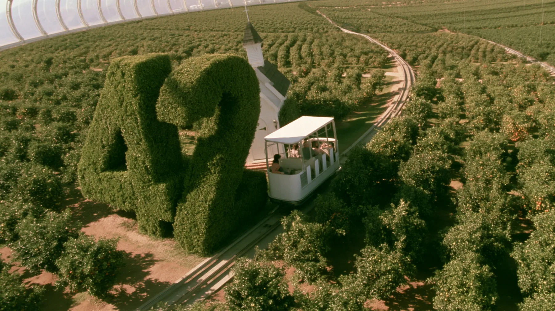 Make this a 35 mm photograph from a small open white tram passing through orchards inside a space colony: beside the track a huge clipped green topiary shaped as the number 42, a small white chapel behind it. Warm…