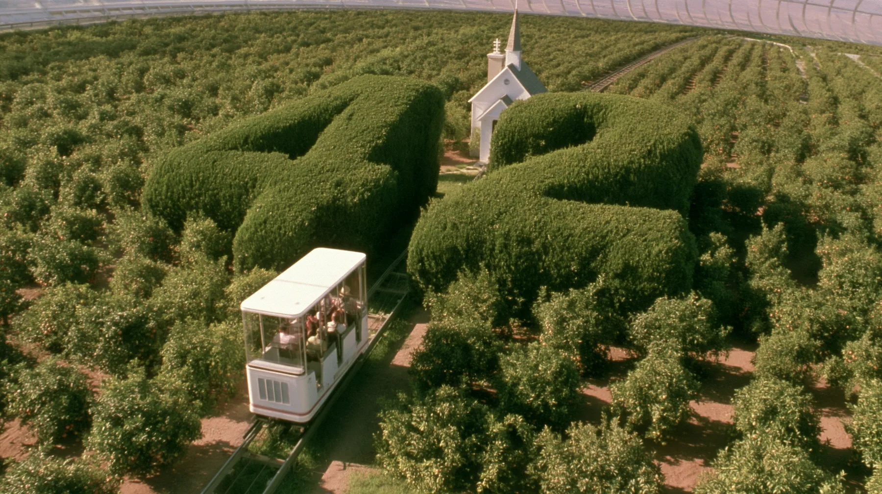 Make this a 35 mm photograph from a small open white tram passing through orchards inside a space colony: beside the track a huge clipped green topiary shaped as the number 42, a small white chapel behind it. Warm…