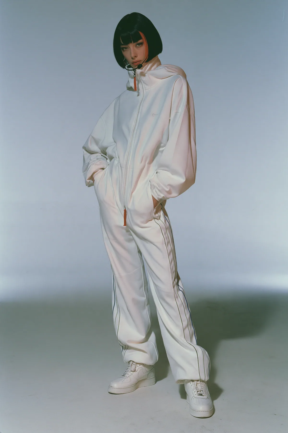 full-length shot, head to toe with the feet in frame, of a young Caucasian American woman with pale skin and a glossy black blunt bob with heavy straight bangs and one clay-orange streak, a thin black headset…