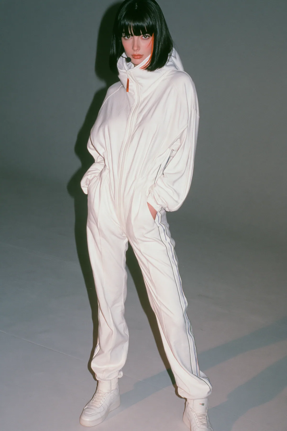 full-length shot, head to toe with the feet in frame, of a young Caucasian American woman with pale skin and a glossy black blunt bob with heavy straight bangs and one clay-orange streak, a thin black headset…