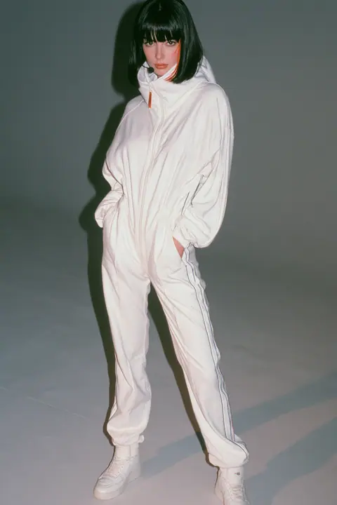 full-length shot, head to toe with the feet in frame, of a young Caucasian American woman with pale skin and a glossy black blunt bob with heavy straight bangs and one clay-orange streak, a thin black headset…