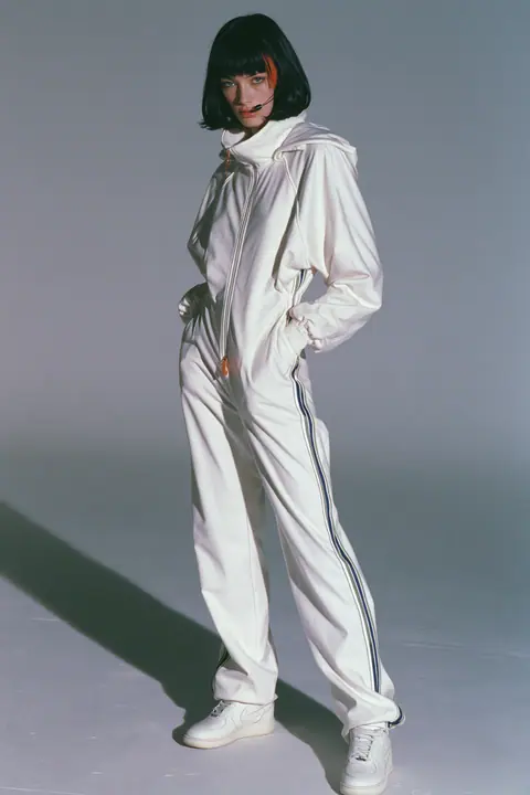 full-length shot, head to toe with the feet in frame, of a young Caucasian American woman with pale skin and a glossy black blunt bob with heavy straight bangs and one clay-orange streak, a thin black headset…