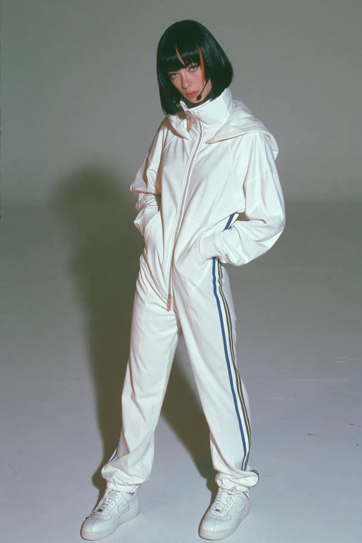 full-length shot, head to toe with the feet in frame, of a young Caucasian American woman with pale skin and a glossy black blunt bob with heavy straight bangs and one clay-orange streak, a thin black headset…