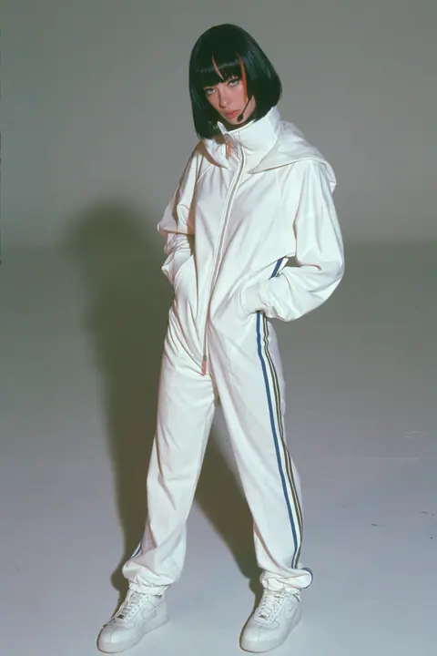 full-length shot, head to toe with the feet in frame, of a young Caucasian American woman with pale skin and a glossy black blunt bob with heavy straight bangs and one clay-orange streak, a thin black headset…
