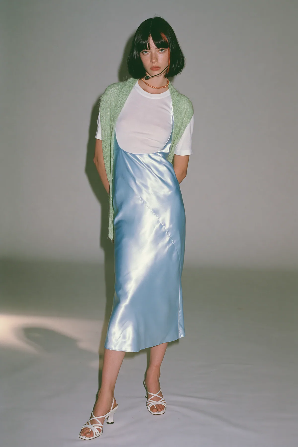 full-length shot, head to toe with the feet in frame, of a young Caucasian American woman with pale skin and a glossy black blunt bob with heavy straight bangs and one clay-orange streak, a thin black headset…